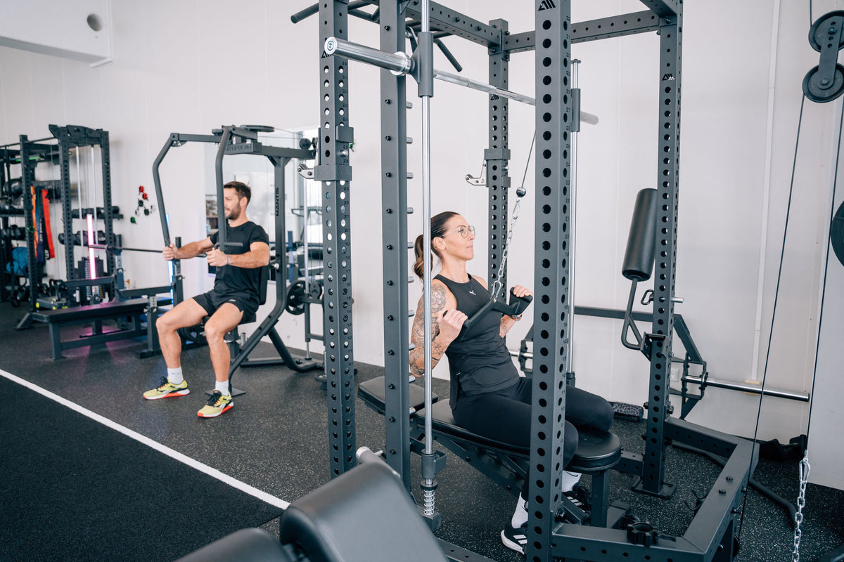 Frau trainiert Latziehen an einem Atletica Power Rack mit Latzug-Anbau, während ein Mann an der Brustpresse im Fitnessstudio trainiert
